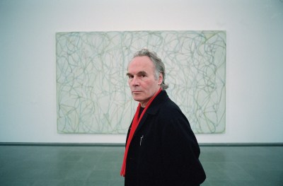 A photograph of a white man in a black jacket and red scarf in front of an abstract painting with squiggles and lines.