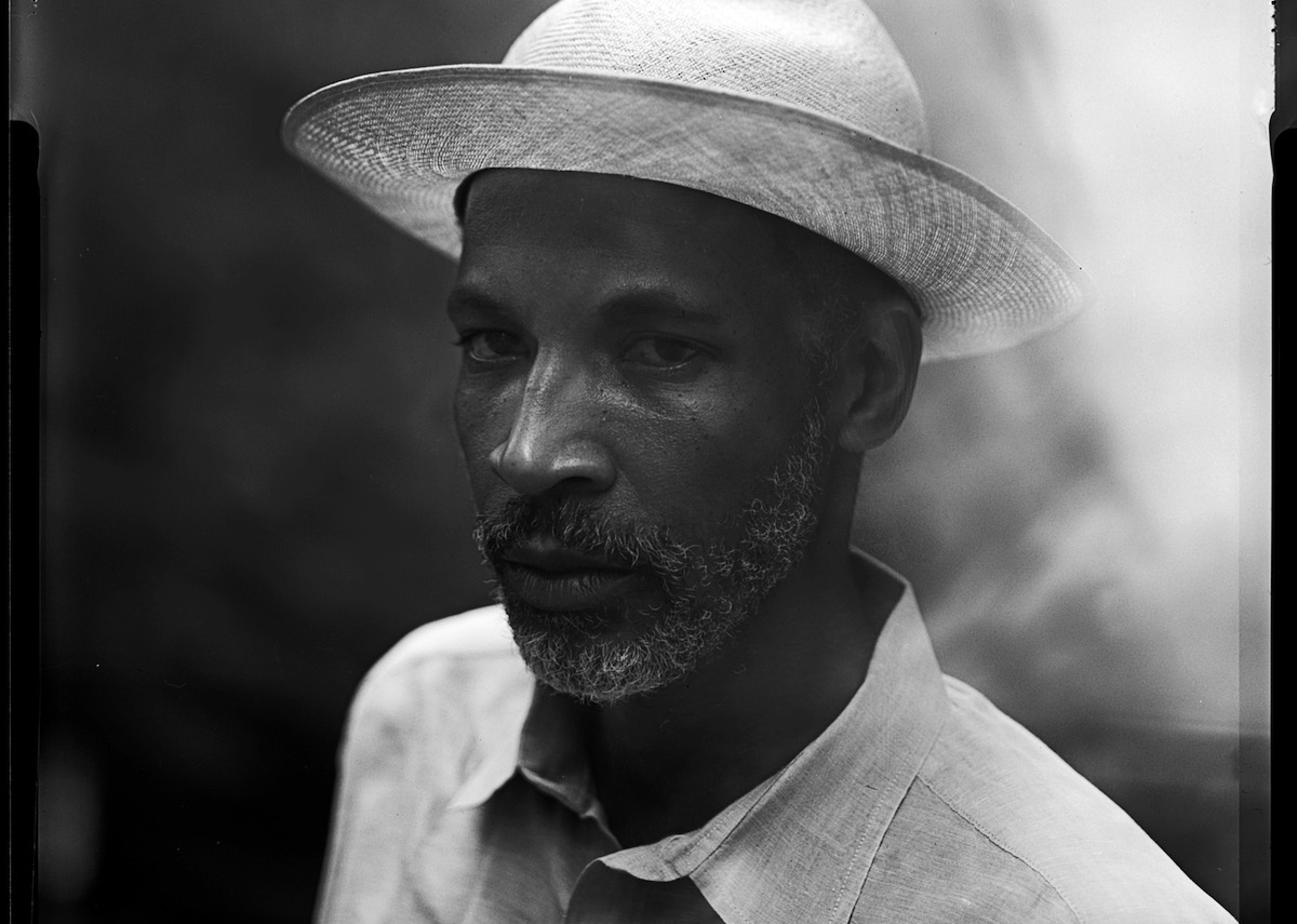 A Black man in a straw hat and a collared shirt looking at the camera.