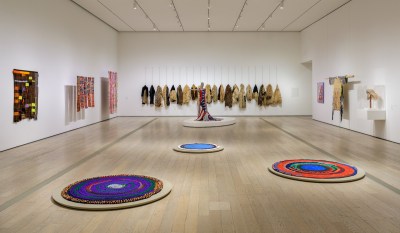 A gallery hung with textiles on its walls, with a few circular ones resembling rippling pools of color on its floors. In the back, a mannequin with a woven dress is on view.