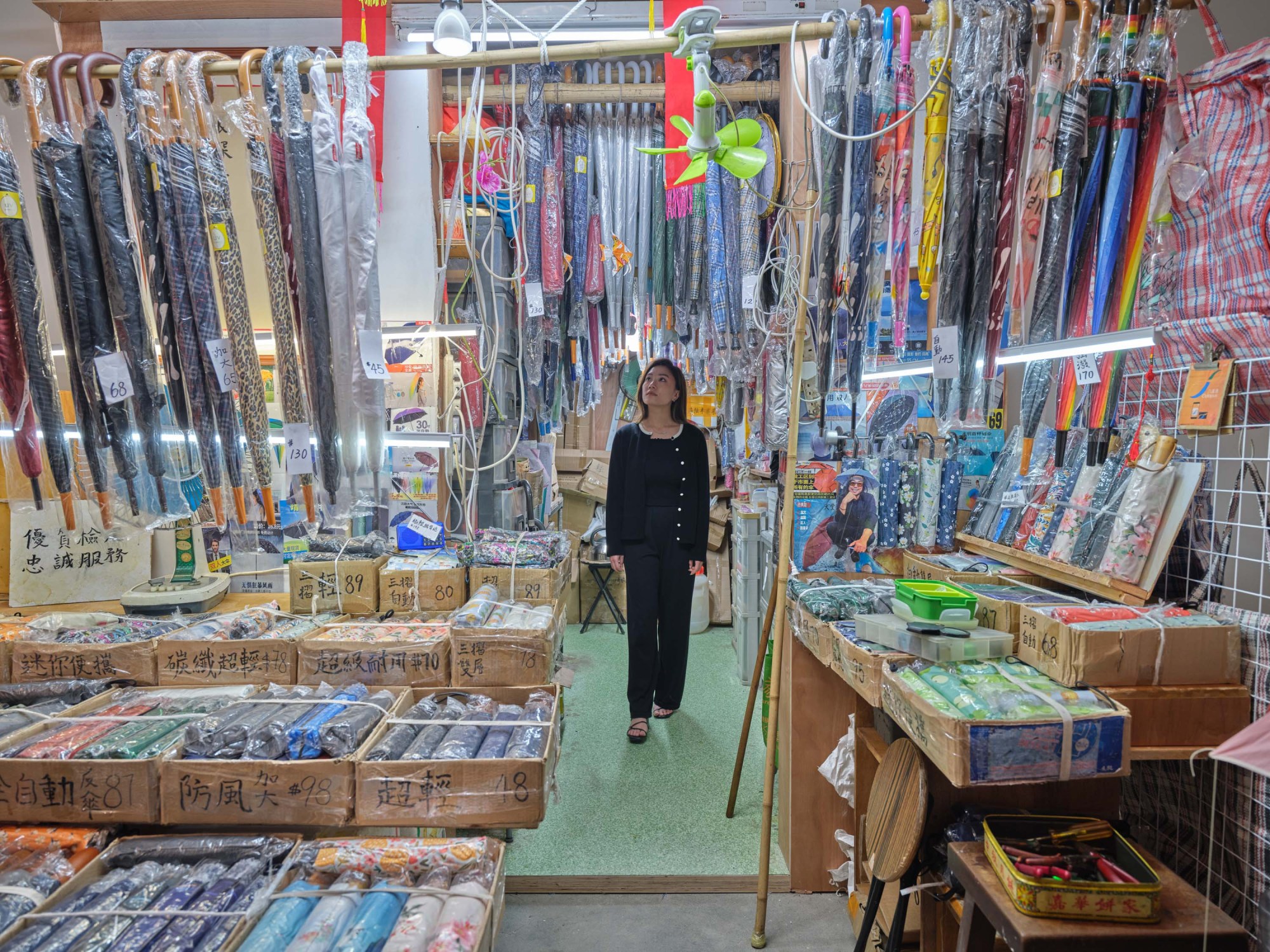 An Asian woman looks up at umbrellas in plastic sleeves in an apparently cluttered and full shop of umbrellas.
