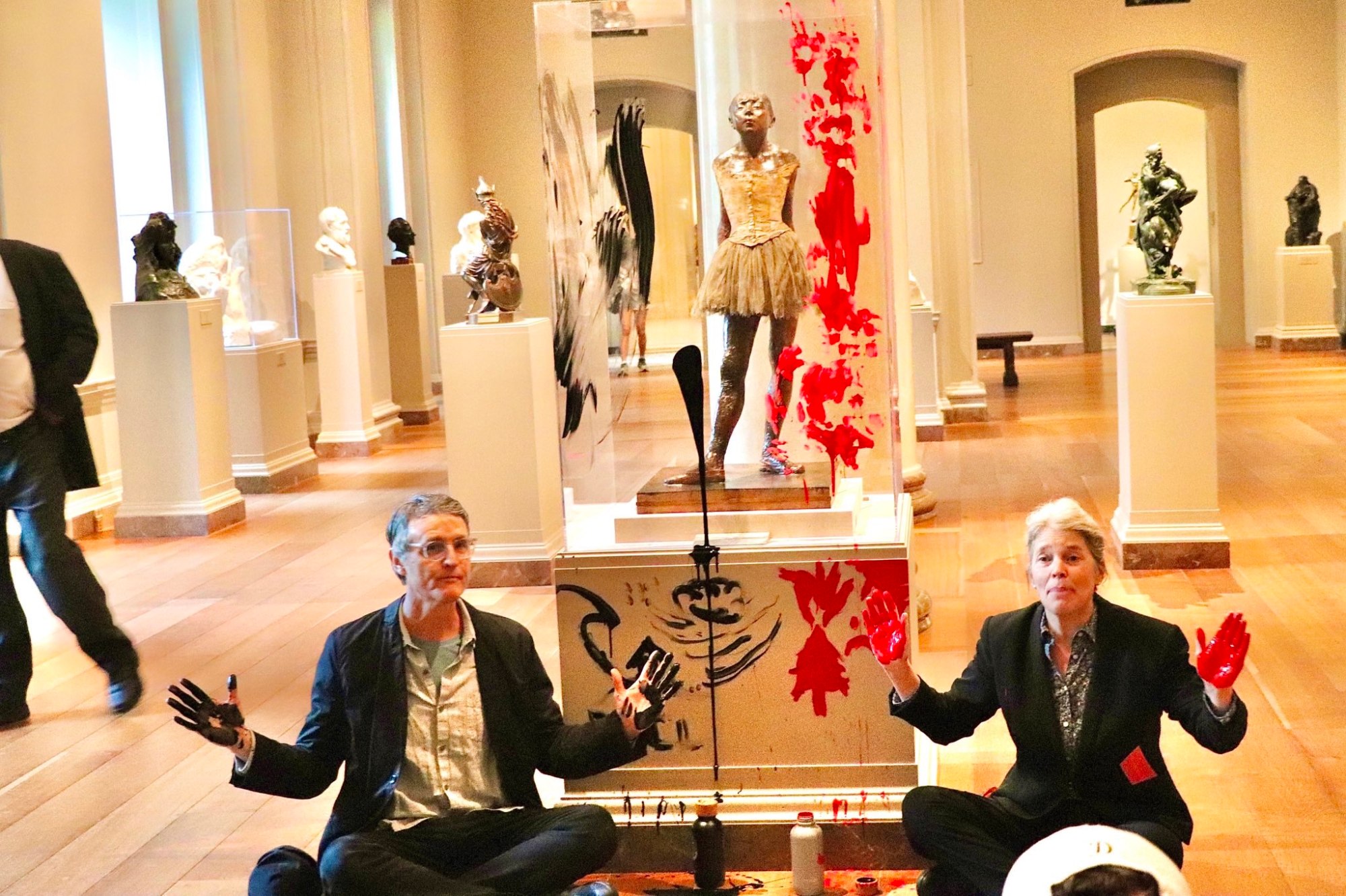 Two climate protesters from the group Declare Emergency are sitting down cross-legged next to the case and pedestal of Degas's sculpture "Little Dancer Aged Fourteen" at the National Gallery of Art after smearing black and red paint on it on April 27, 2023. The protestors' hands are covered in paint.