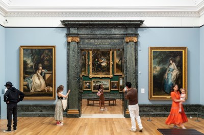 A group of contemporary museum visitors looking at a gallery hung with tall portraits of centuries-old sitters. Through a hallway is a view of a more densely hung gallery.