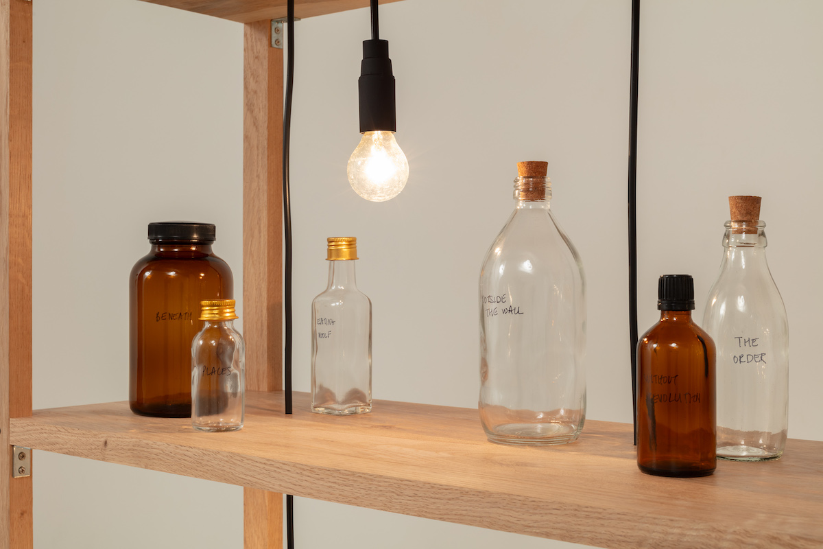 Six glass bottles on a wooden shelf, with a lightbulb hanging above.