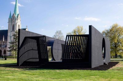 A large black sculpture with tilted areas composed of bars. The sculpture is set in a green area with a church in the background.