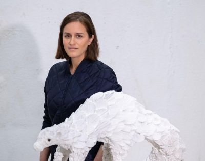 A woman with olive skin and caramel hair stands next to a styrofoam sculpture of a pangolin.