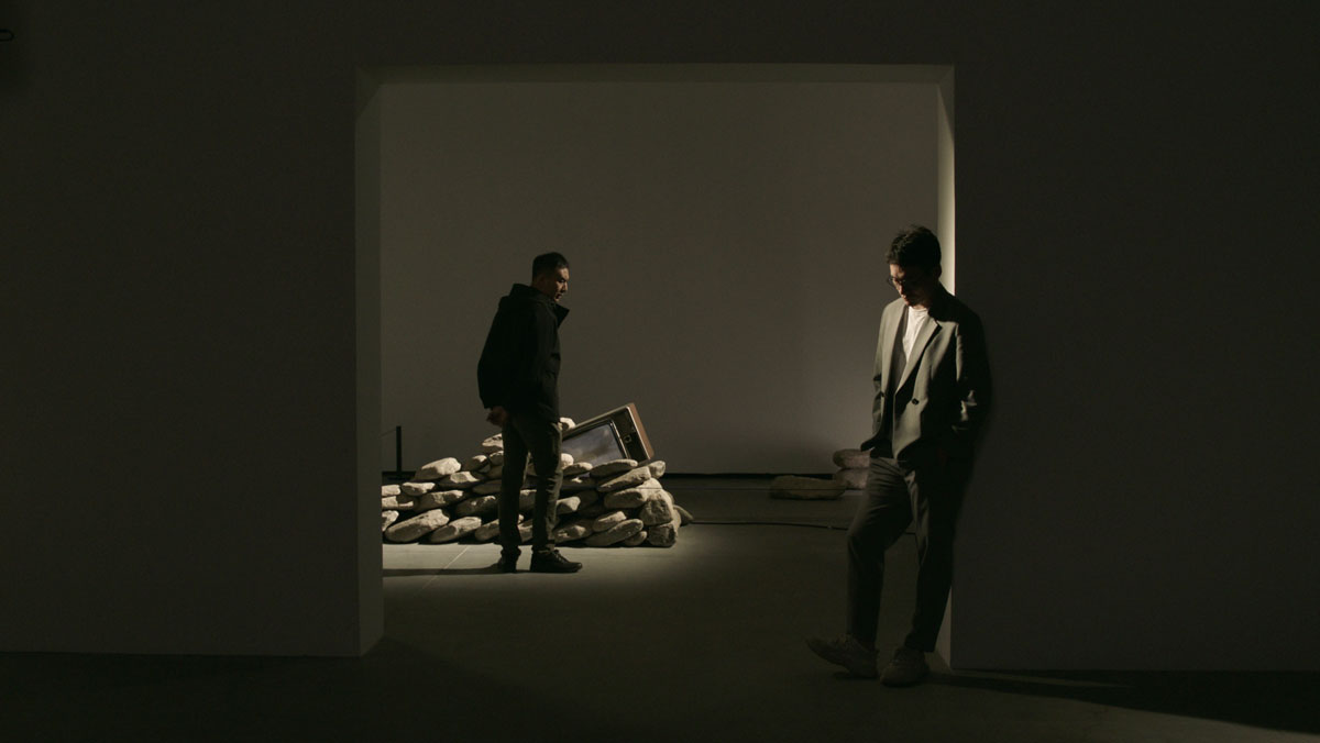 A film still showing two men in a dimly lit art gallery. One, who stands before an art installation, looks back at the other, who stands looking down in an entryway.
