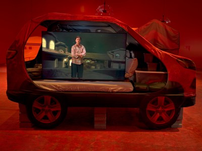 A video screen showing a man with his arms folded inside a sculpture of a van with a bed inside.