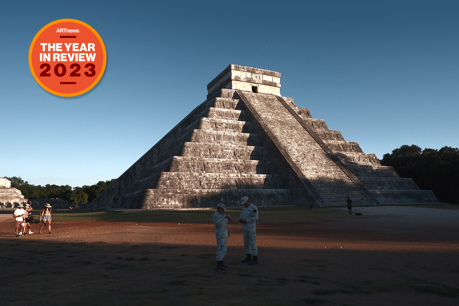The pyramid complex of Chichen Itza is seen near sunset with tourists standing in front.