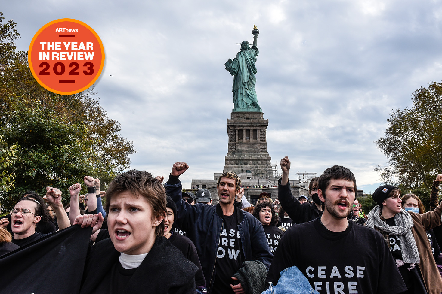 Many people in black shirts reading "Ceasefire Now" shout in front of the Statue of Liberty.