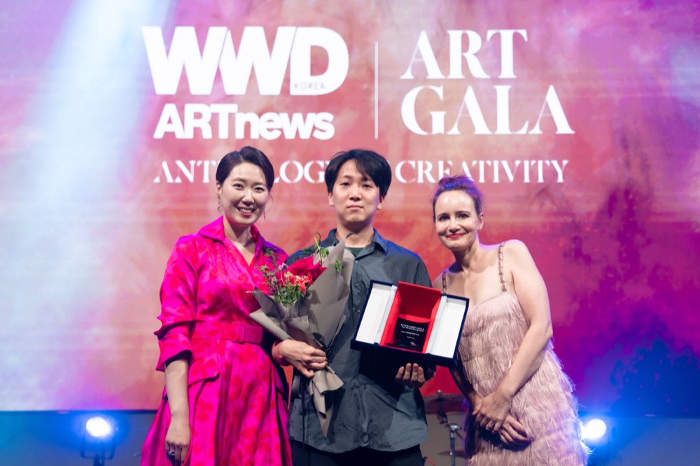 An Asian woman in a magenta dress, an Asian man in dark gray shirt, and a Caucasian woman in a light pink dress stand in front of a backdrop that reads, "WWD ARTnews Art Gala."