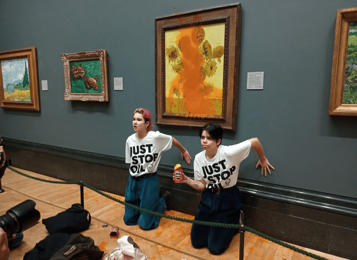 Two white protesters in tee shirts that say "just stop oil" have their hands glued to the wall in front of Van Gogh's sunflowers, which is covered in tomato soup.